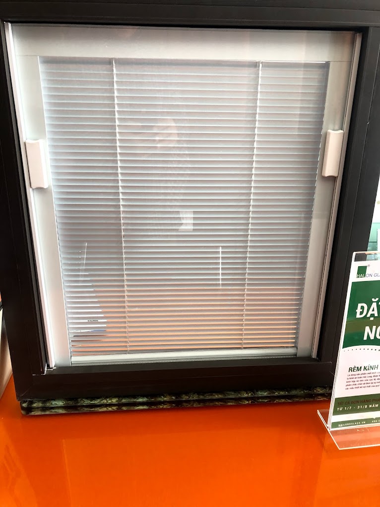 Black aluminum-framed windows with integrated horizontal blinds inside the glass display case on an orange tabletop.