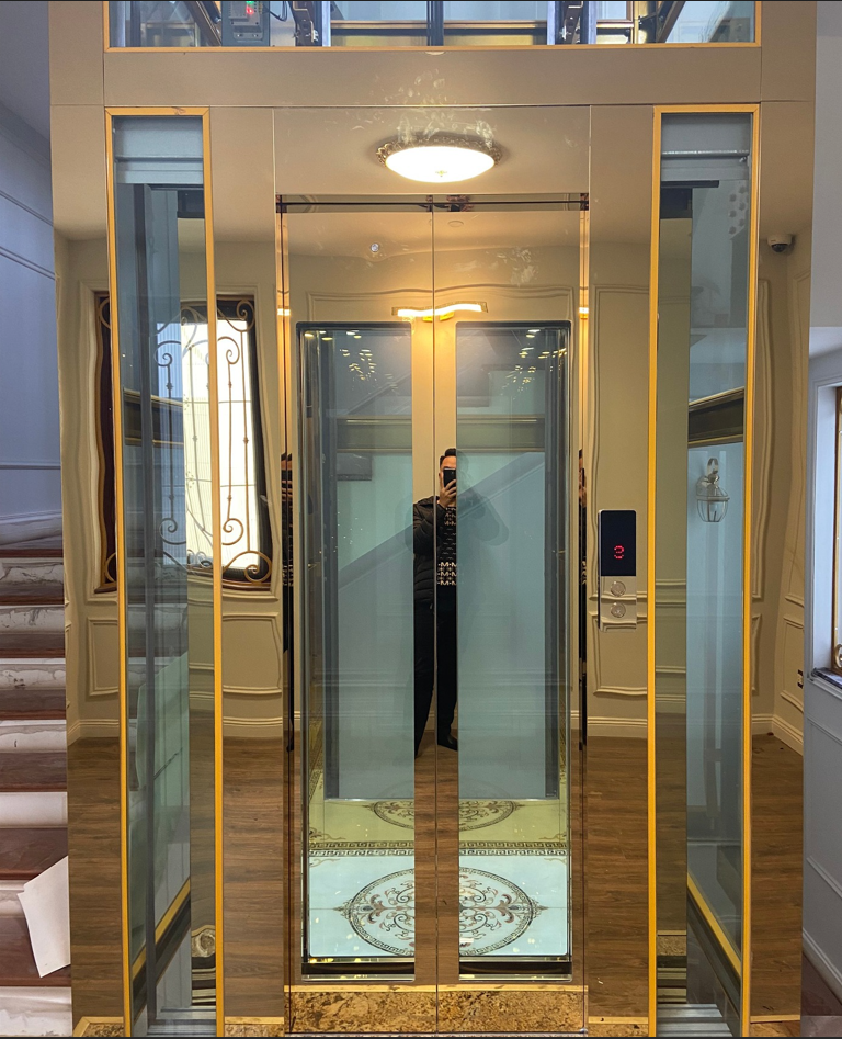 A luxurious gold-framed glass elevator inside the building, with glass doors reflecting the interior and a wooden staircase beside it.