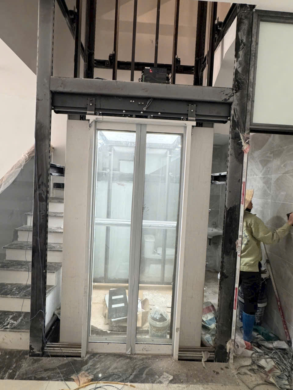 A glass-doored home elevator is being installed between two stairwells in a newly constructed house.