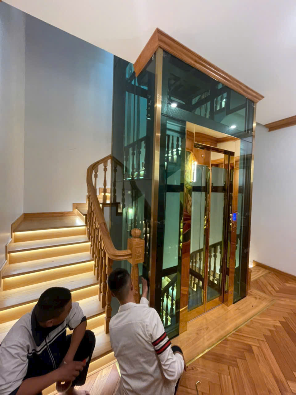A glass-framed, wood-paneled home elevator is being installed by technicians next to a wooden staircase with LED lighting.