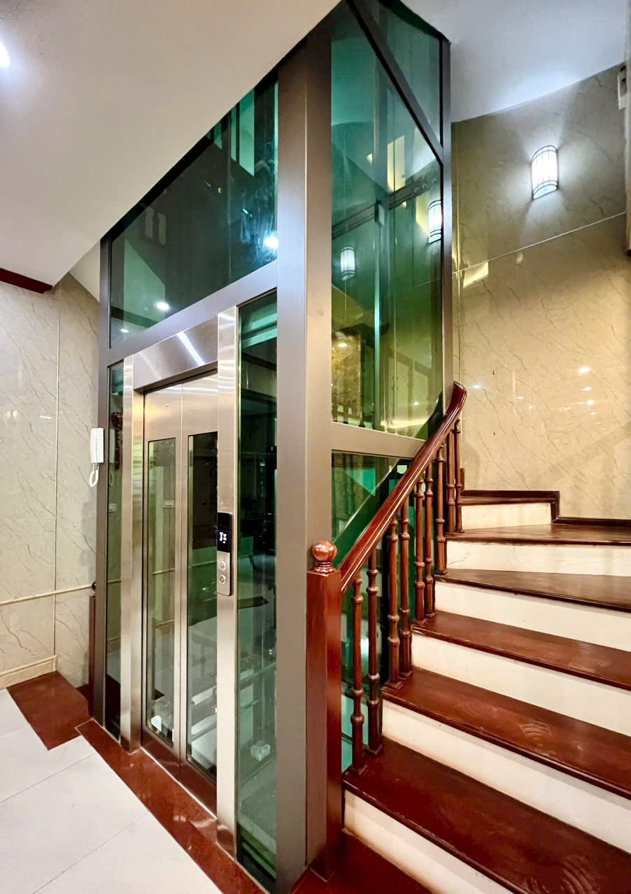 Glass-walled home elevator next to a wooden staircase in a luxurious house.